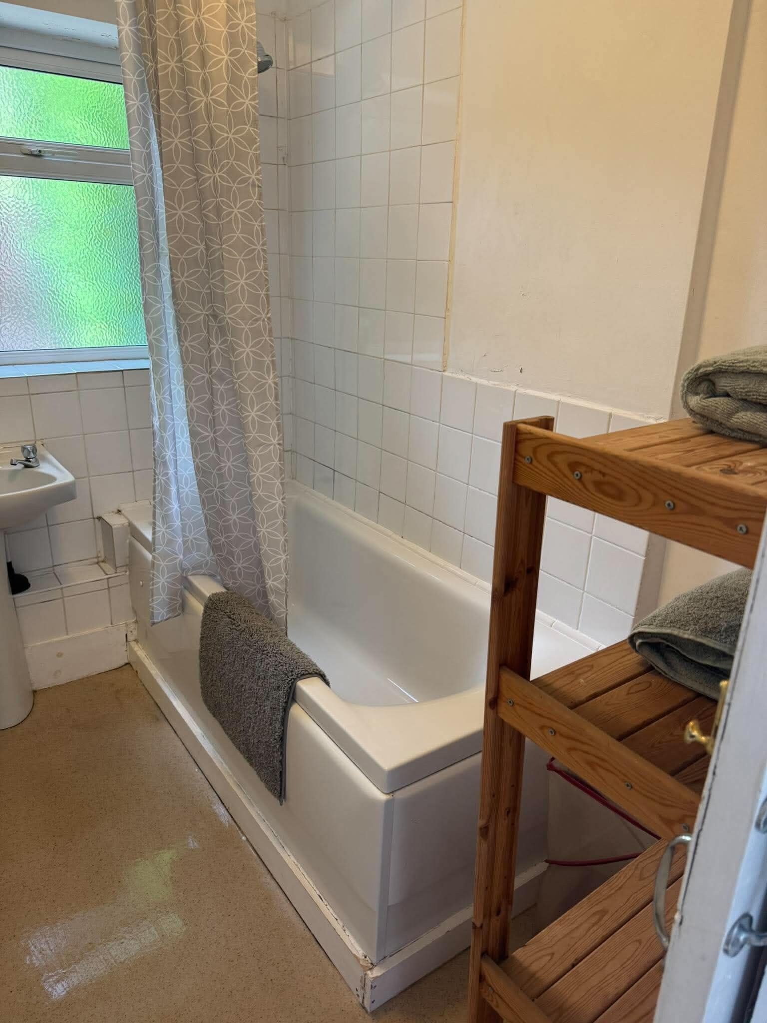 A bathroom with a white bathtub, a wooden shelf, and a window with a patterned curtain.