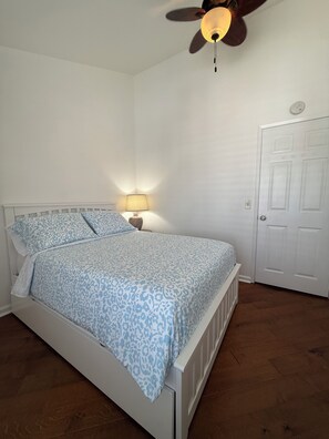 A bedroom with a bed, a ceiling fan, a nightstand with a lamp, and a door. - Home Away By the Beach 2! Beautiful 3-bedroom Newport Beach Upstairs Condo (Newport Beach)
