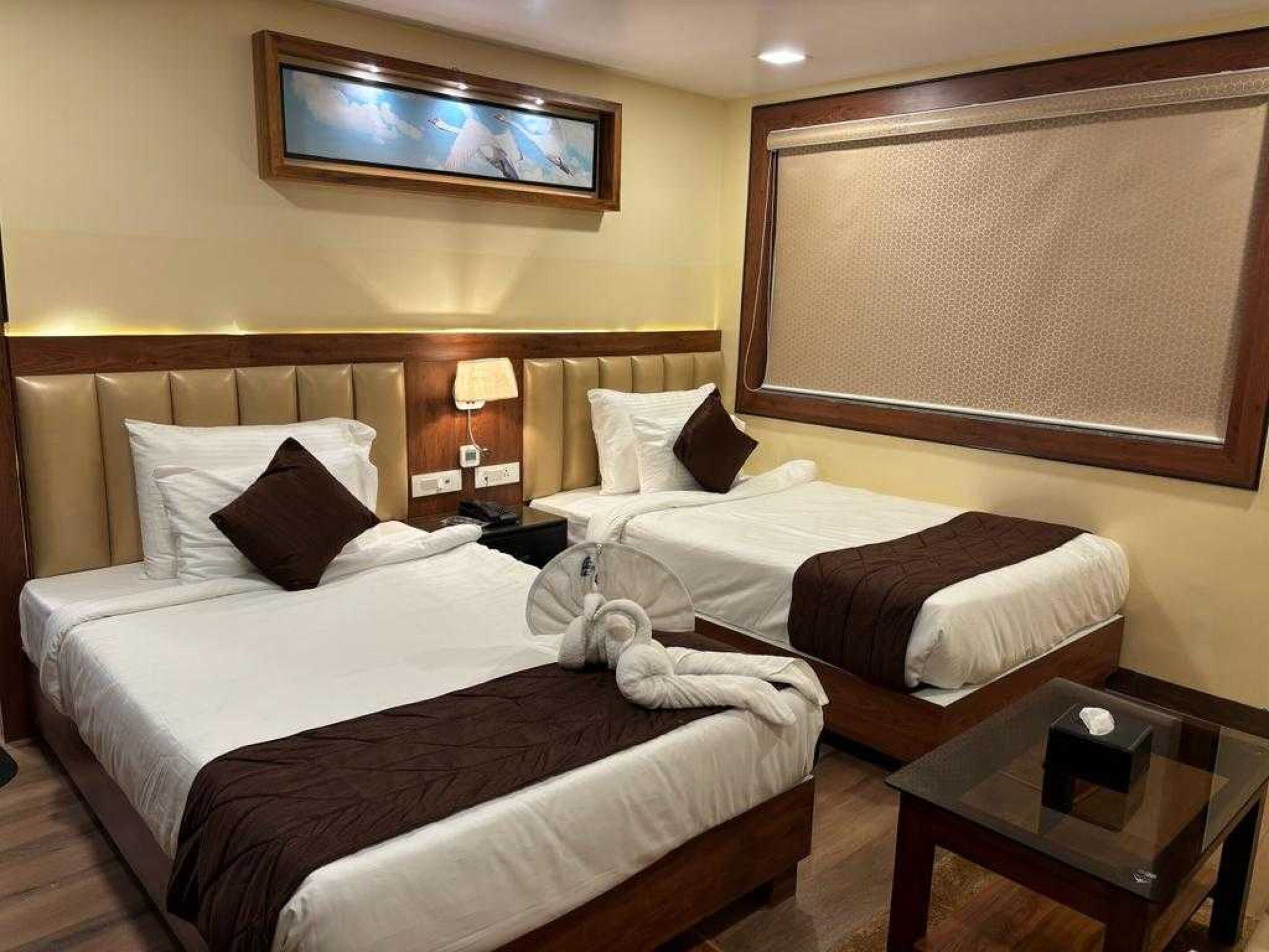 A hotel room with two beds, a bedside table, a wall-mounted TV, and a large window.