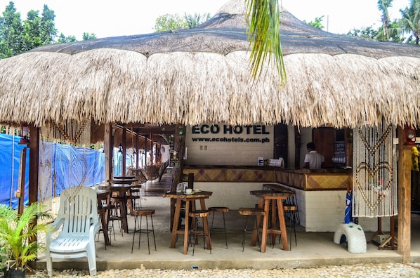 Un área de estar al aire libre con techo de paja, mesas y sillas de madera, y un letrero que dice ECO HOTEL.