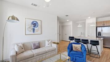 Una sala de estar moderna con un sofá, un sillón azul y una mesita de centro. Hay una zona de cocina con un refrigerador y una barra.