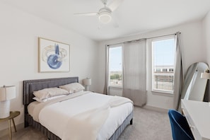 Ein Hotelzimmer mit einem Bett, einem Schreibtisch, einem Stuhl, einem Deckenventilator und einem fenster mit Vorhängen.