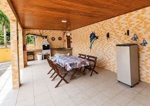 Een terras buiten, met een bakstenen muur, houten afdak, betegelde vloer, een tafel met stoelen, een koelkast en een barbecue.