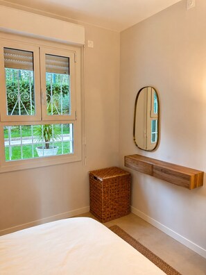 Ein Schlafzimmer mit einem Bett, einem Fenster mit Jalousien, einem Spiegel, einer kleinen Holzkiste und einem Regal.