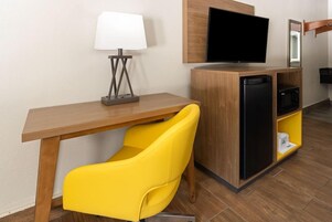 Habitación de hotel con un escritorio de madera, una silla amarilla, un televisor de pantalla plana, un refrigerador y un microondas.