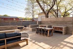 Een terras buiten met een houten bank, een tafel en stoelen, omgeven door een houten hek en bomen.