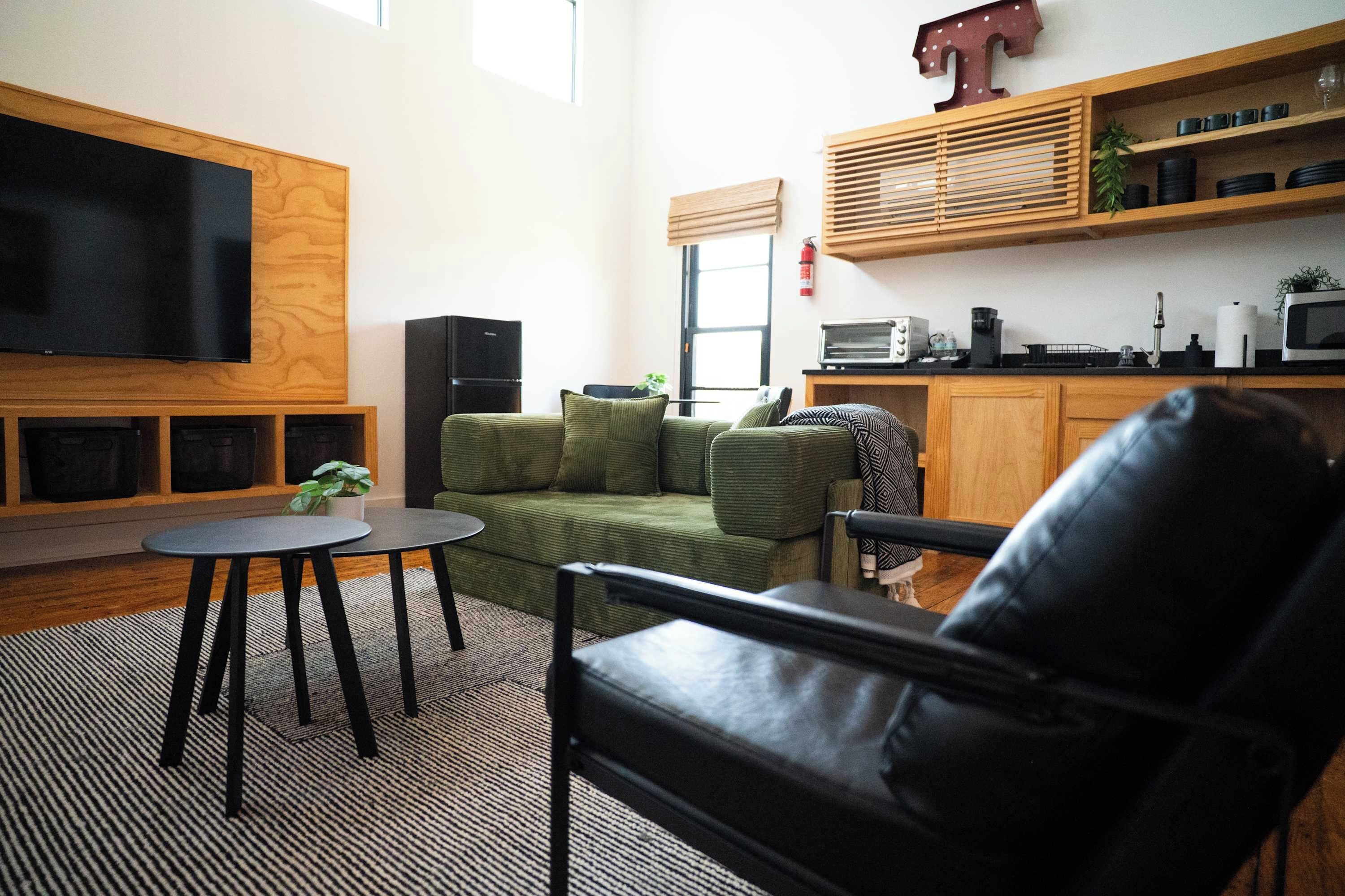 Ein Wohnzimmer mit einem grünen Sofa, einem schwarzen Ledersessel, einem kleinen runden Tisch und einem an der Wand befestigten Flachbildfernseher.