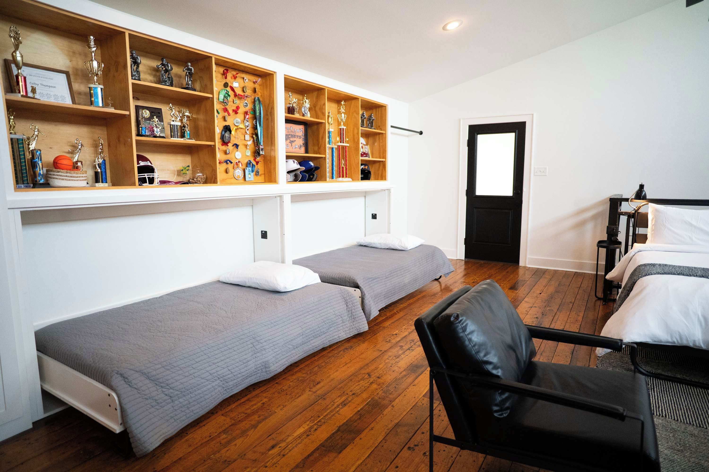 Ein Zimmer mit zwei Einzelbetten, einem Holzboden mit Pokalen und Medaillen, einem schwarzen Ledersessel und einer Tür.