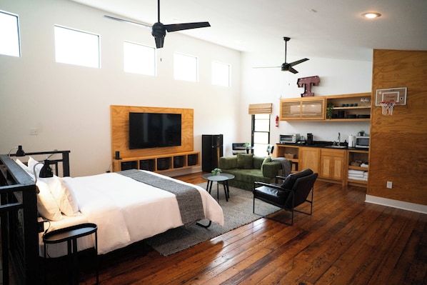 Quarto de hotel moderno com uma cama grande, área de estar, cozinha compacta e televisão na parede.