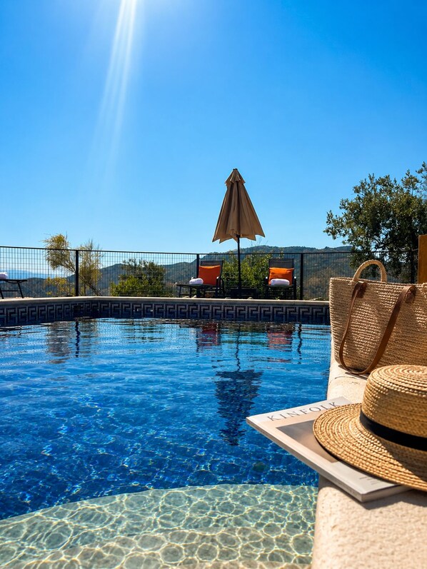 Een helderblauw zwembad met een zonnige lucht, een loungegedeelte met stoelen en een parasol, en een geweven mand aan de rand.