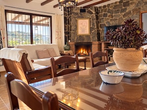A cozy living room with a stone fireplace, a sofa, a dining table with chairs, and a large window with curtains. - Country House with a Jacuzzi, Pool, and Spectacular Mountain Views (Colmenar)