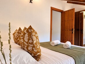 Een net opgemaakt bed met witte lakens en een bruin gebloemd kussen, een groene plaid en twee opgerolde handdoeken.