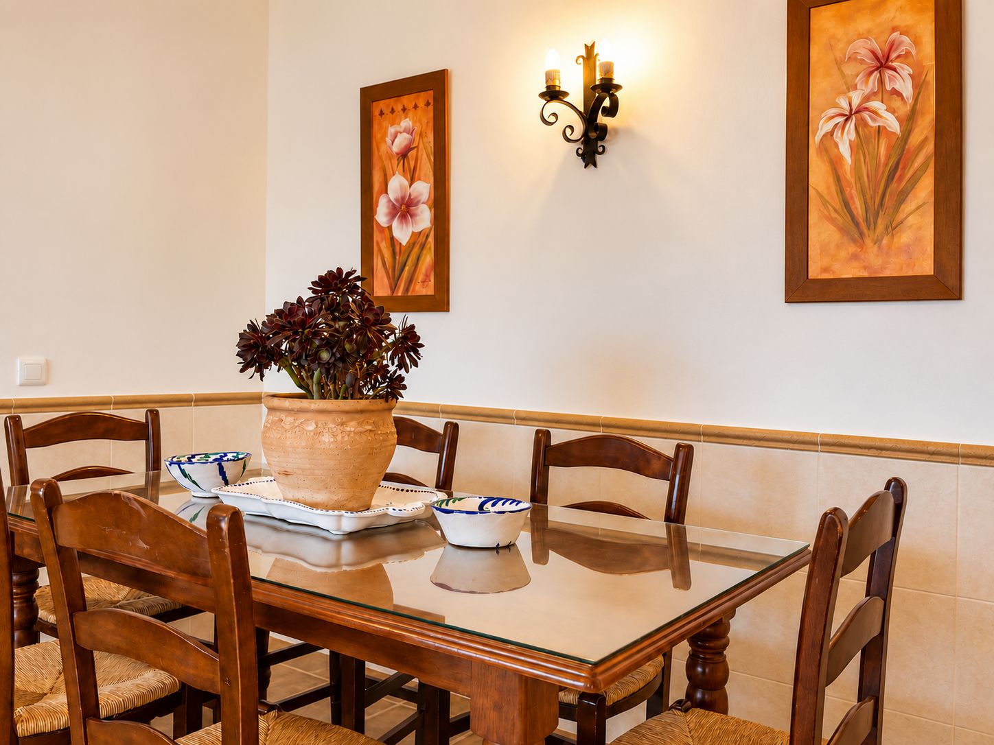 A dining area with a glass-top table, wooden chairs, a potted plant, and wall-mounted lighting.