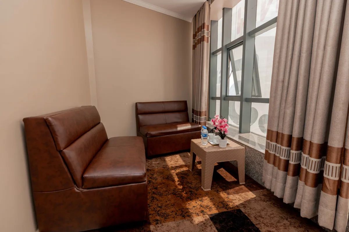 Una habitación con un sofá de cuero marrón, un sillón más pequeño, una mesita con un jarrón de flores y una ventana con cortinas.