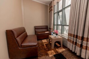 A room with a brown leather sofa, a smaller armchair, a small table with a vase of flowers, and a window with curtains. - Gihozo Hotel (Kigali)