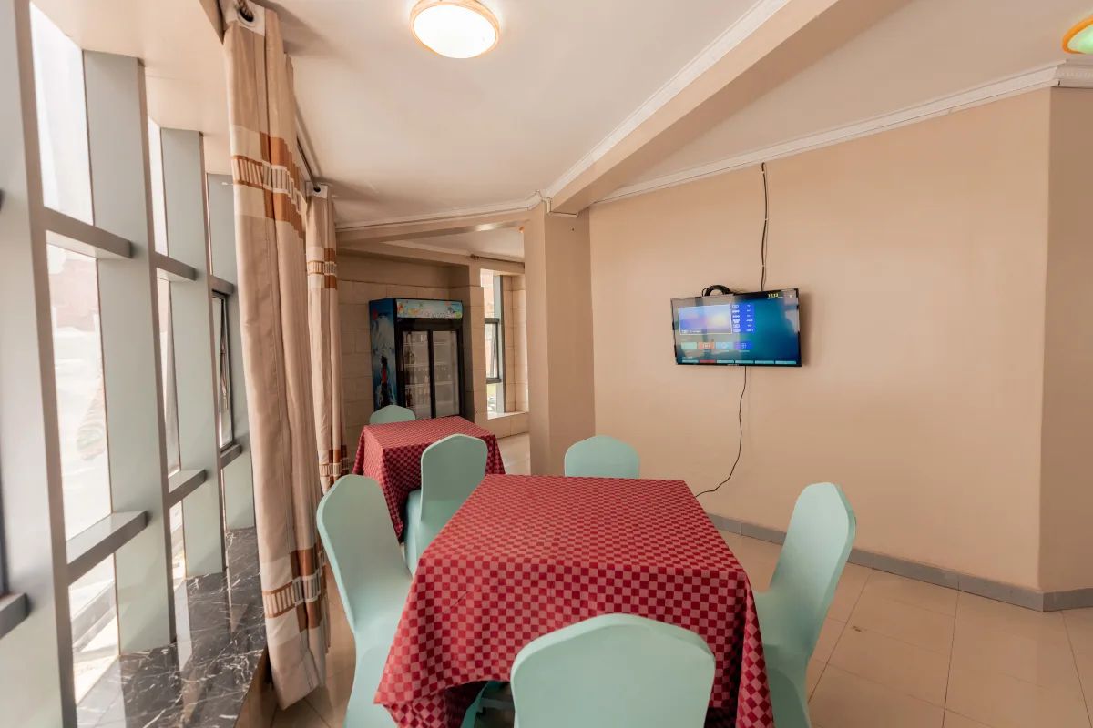 A hotel dining area with tables and chairs, a television mounted on the wall, and large windows with curtains.