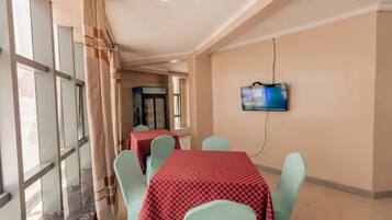 A hotel dining area with tables and chairs, a television mounted on the wall, and large windows with curtains.