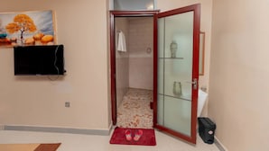 A bathroom with a glass door, a shower, a tiled floor, a red mat, a trash bin, and a television mounted on the wall. - Gihozo Hotel (Kigali)