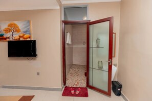 A bathroom with a glass door, a shower, a tiled floor, a red mat, a trash bin, and a television mounted on the wall. - Gihozo Hotel (Kigali)