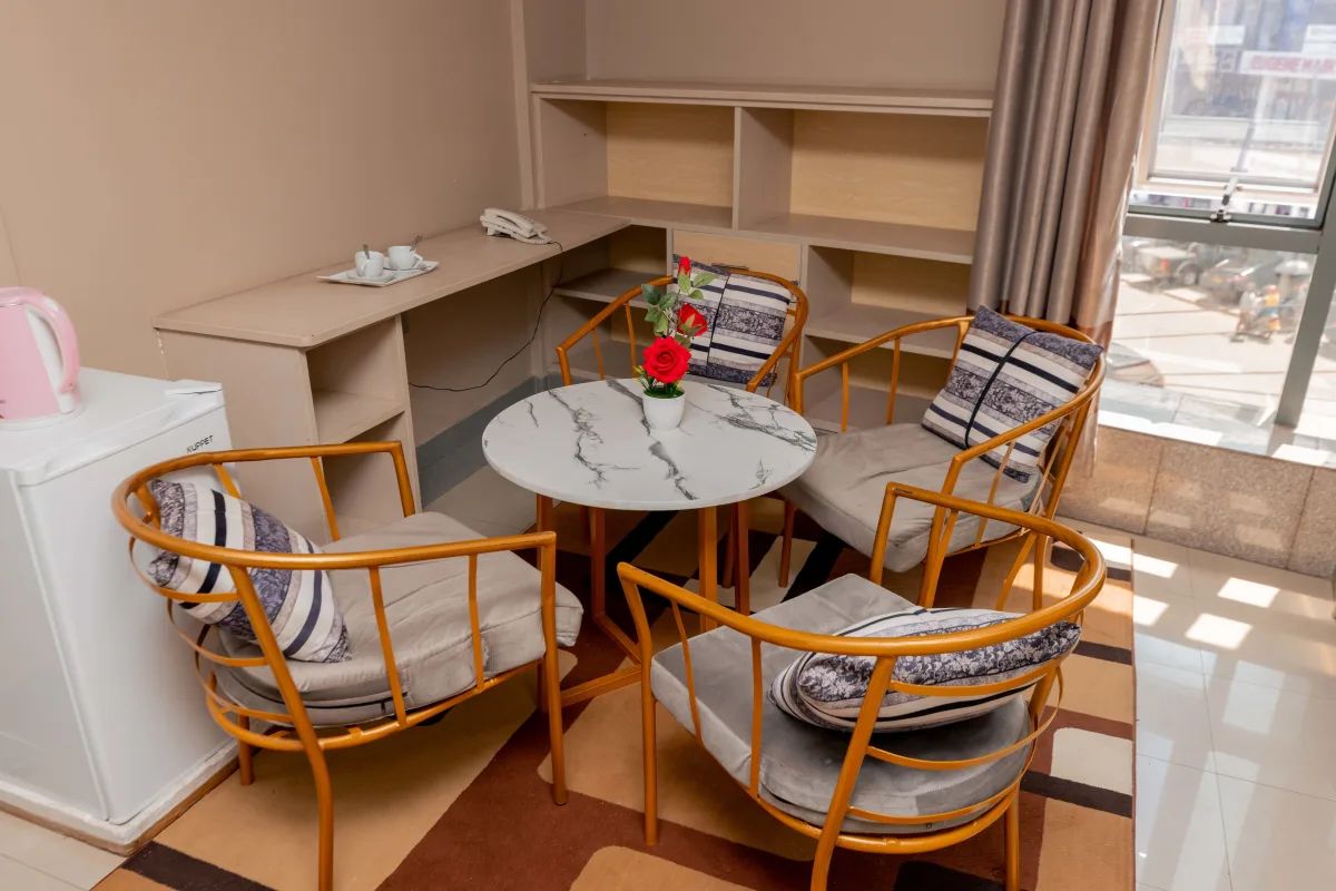 Quarto com uma mesa redonda, quatro cadeiras, um pequeno vaso com flores, uma chaleira e um telefone em uma prateleira.