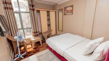 Een hotelkamer met een bed, een bureau, een stoel, een ventilator en een raam met gordijnen.