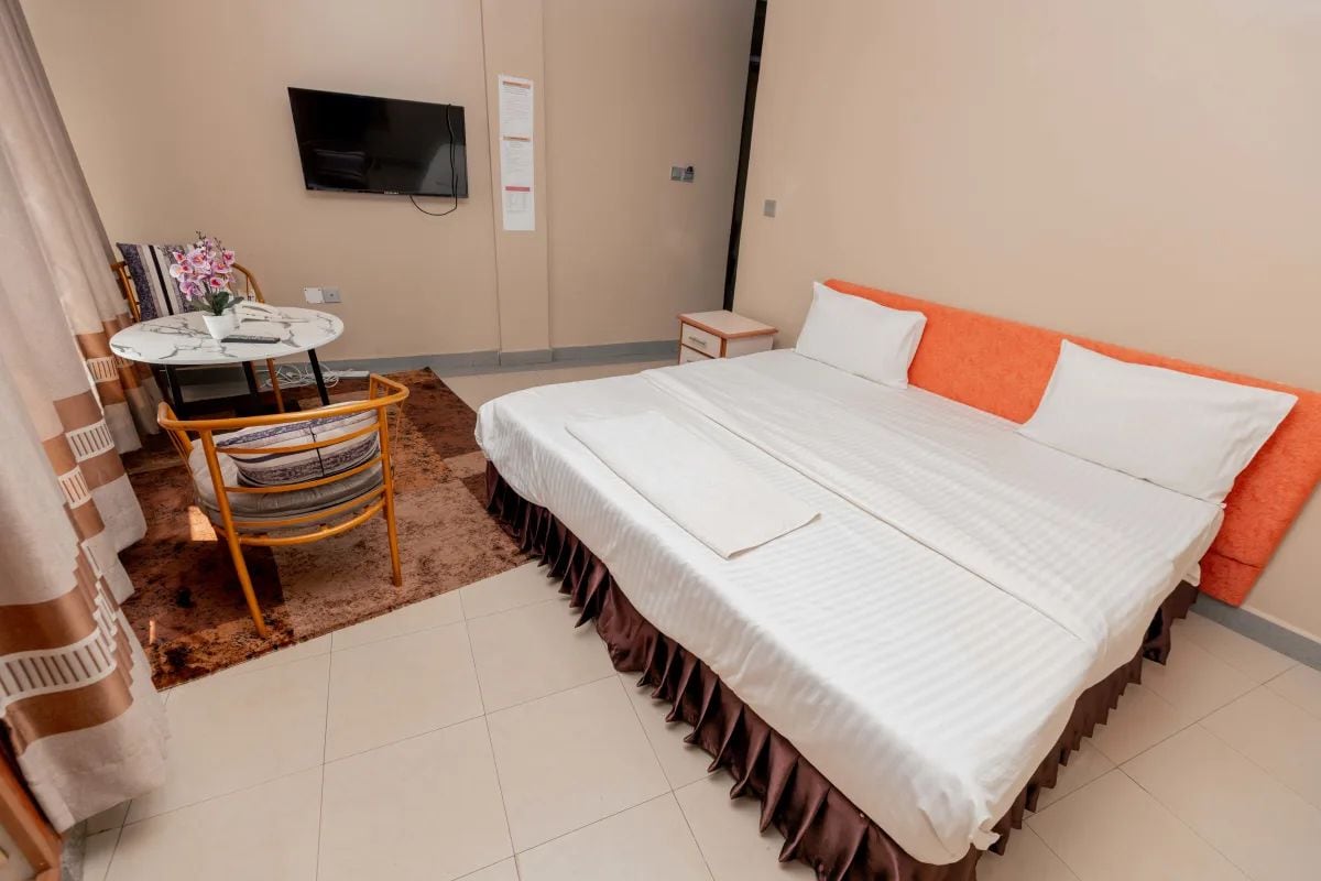 Quarto de hotel com cama, televisão fixada na parede, mesa pequena com cadeira e tapete.