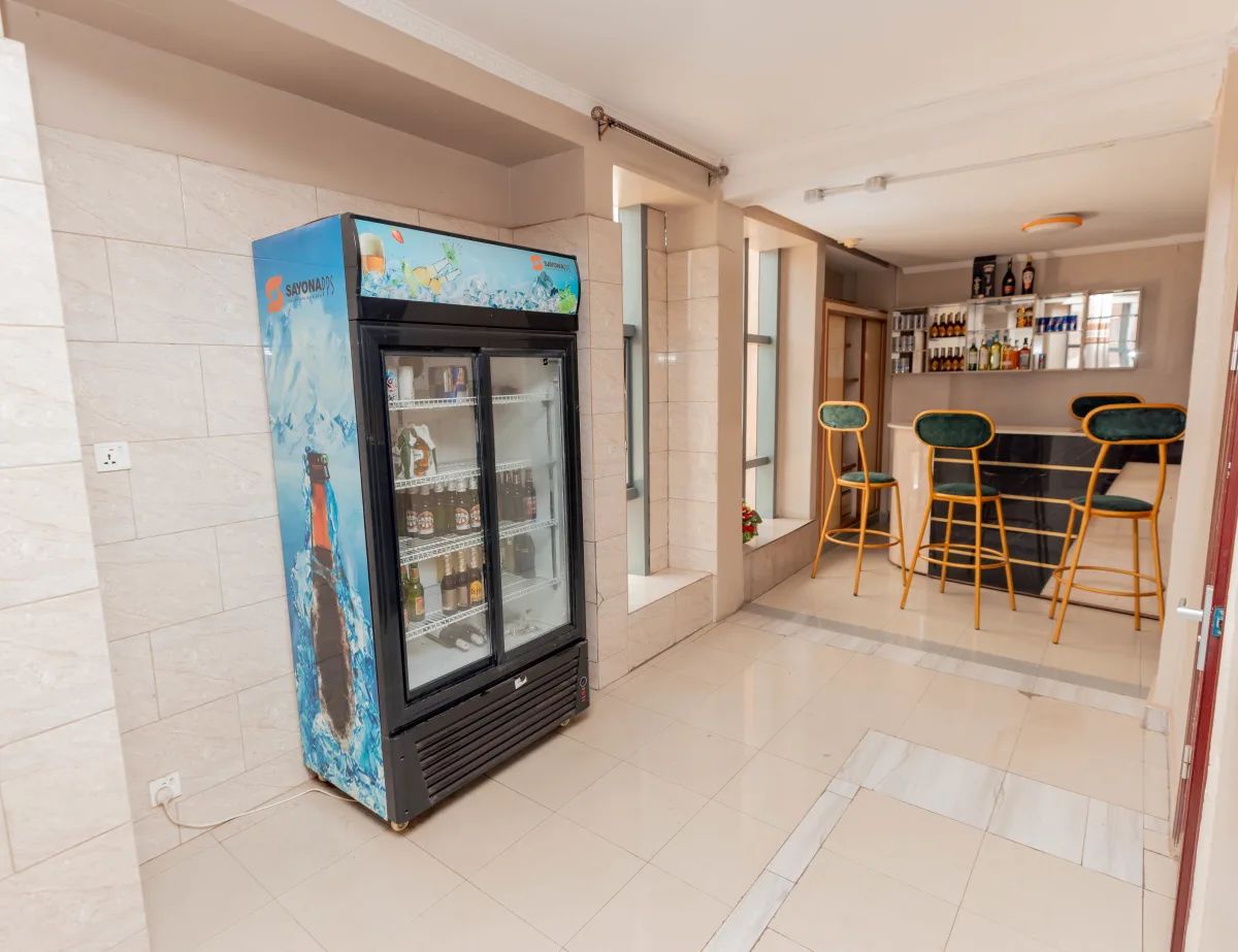 Um saguão de hotel com minibar, área de bar com banquetas e exposição de bebidas engarrafadas.