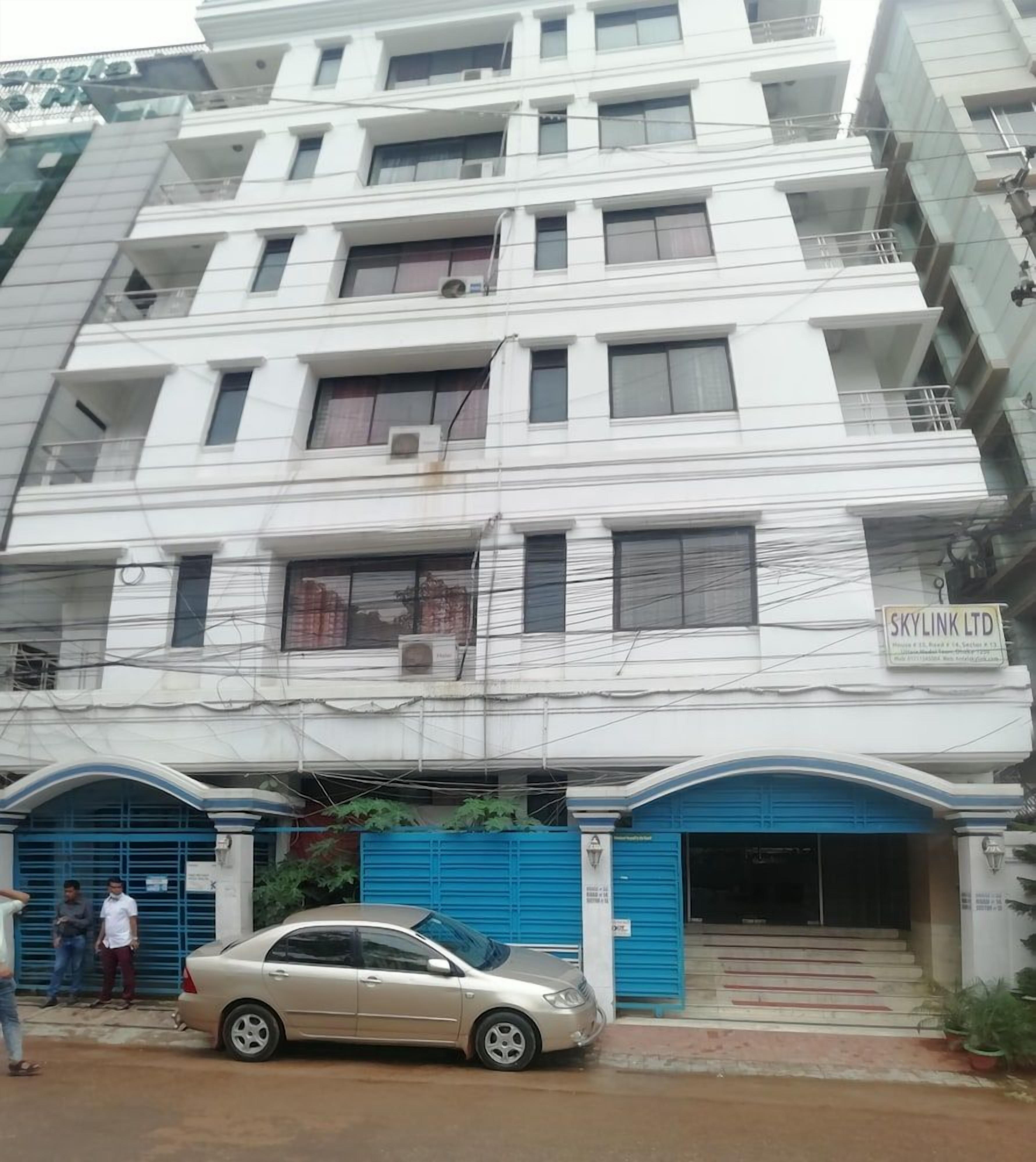 A multi-story building with a blue entrance gate, a car parked in front, and a sign for 'SKYLINK LTD' on the facade.