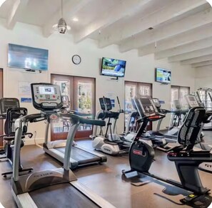 Ein Fitnessraum mit mehreren Laufbändern und Trainingsgeräten, einer Uhr an der Wand und einem Fernseher, der darüber angebracht ist.
