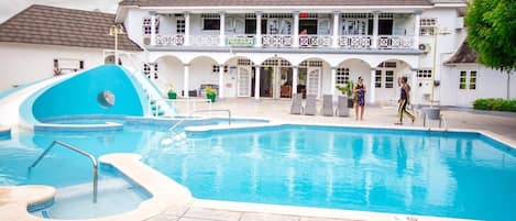 Uma piscina com escorrega, um edifício com várias varandas e algumas pessoas.