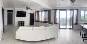 A modern living room with a white sectional sofa, a flat-screen TV mounted on the wall, a ceiling fan, and large windows with curtains. - Mountain Oasis-Farm Stay solar powered with swimming pool. Quiet and peaceful. (San German)