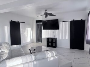 A modern living room with a white sofa, a coffee table, a ceiling fan, and a flat-screen TV mounted on the wall. - Mountain Oasis-Farm Stay solar powered with swimming pool. Quiet and peaceful. (San German)