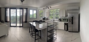 A modern kitchen with a central island, bar stools, a refrigerator, and a view of greenery through large windows. - Mountain Oasis-Farm Stay solar powered with swimming pool. Quiet and peaceful. (San German)