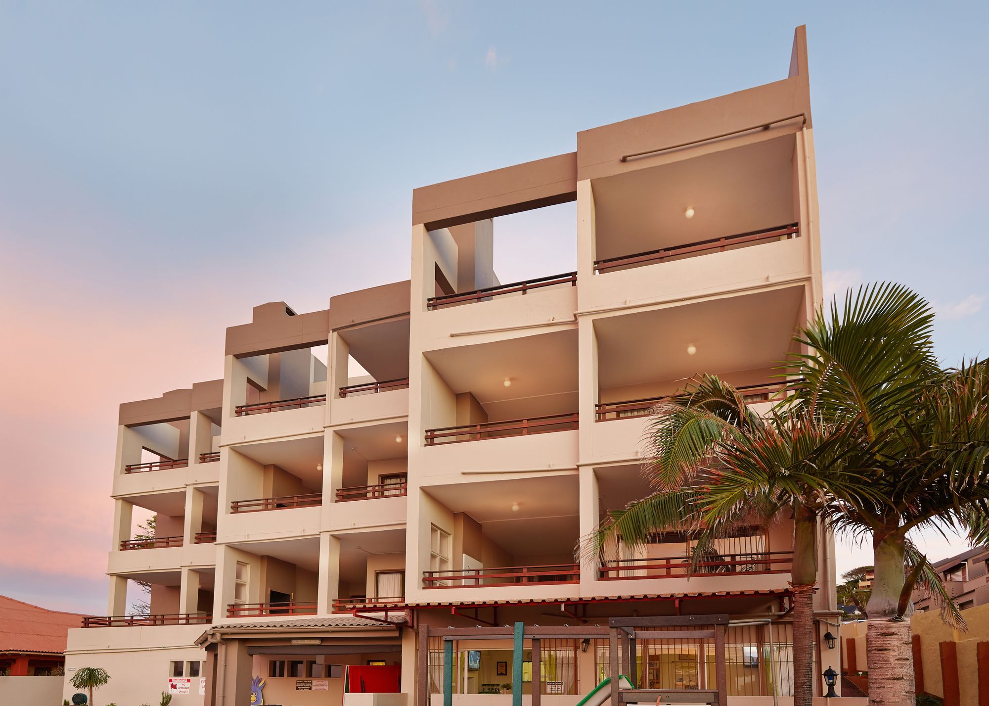 A multi-story hotel with balconies, a covered entrance, and a palm tree.
