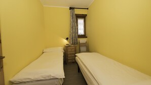 Een smalle kamer met twee enkele bedden, een houten dressoir, een raam met gordijn en een radiator aan de muur.