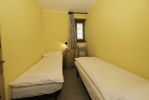 Habitación pequeña con dos camas individuales, un aparador de madera, una ventana con cortina y un radiador fijado a la pared.