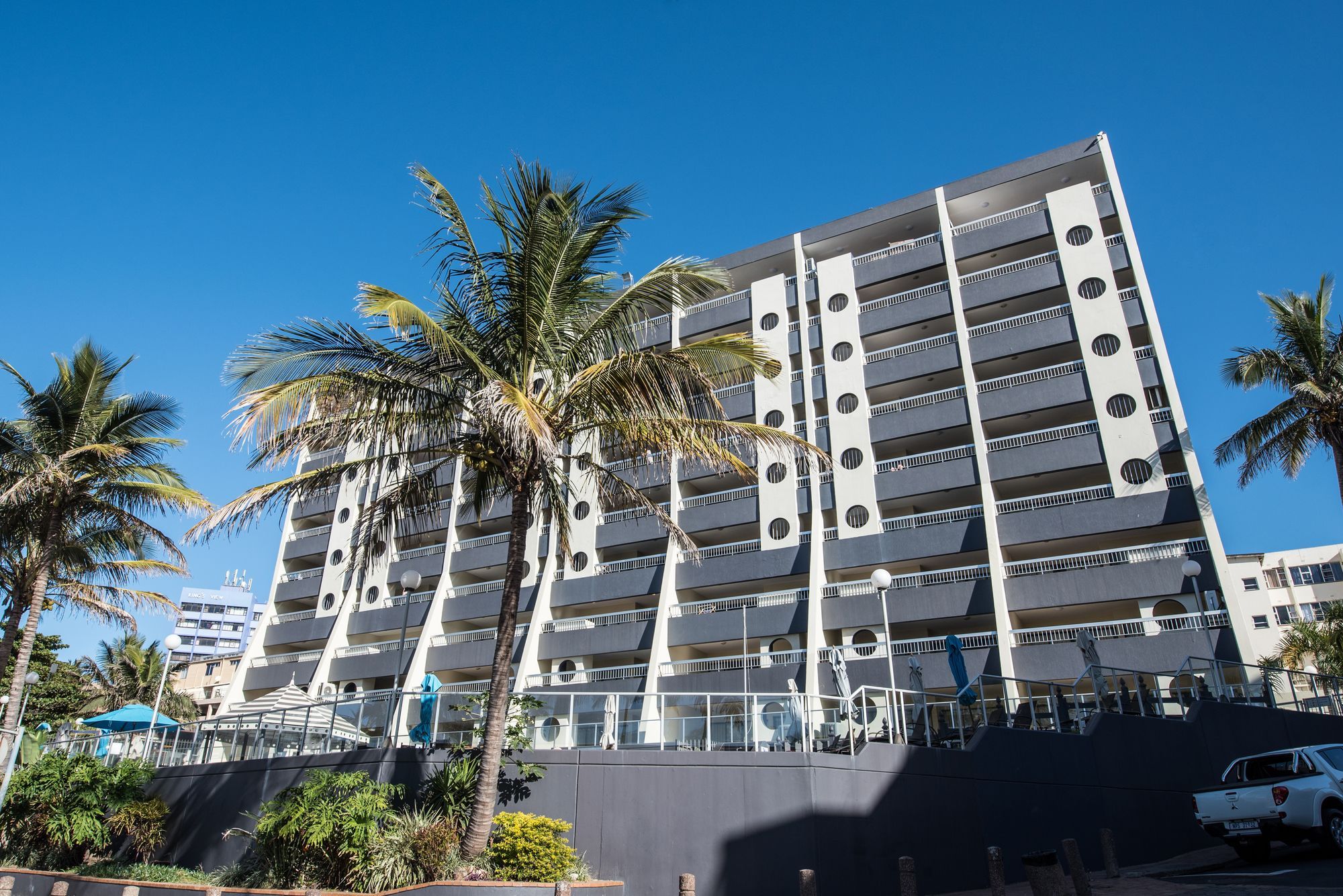 A multi-story building with balconies, palm trees, and a clear blue sky.