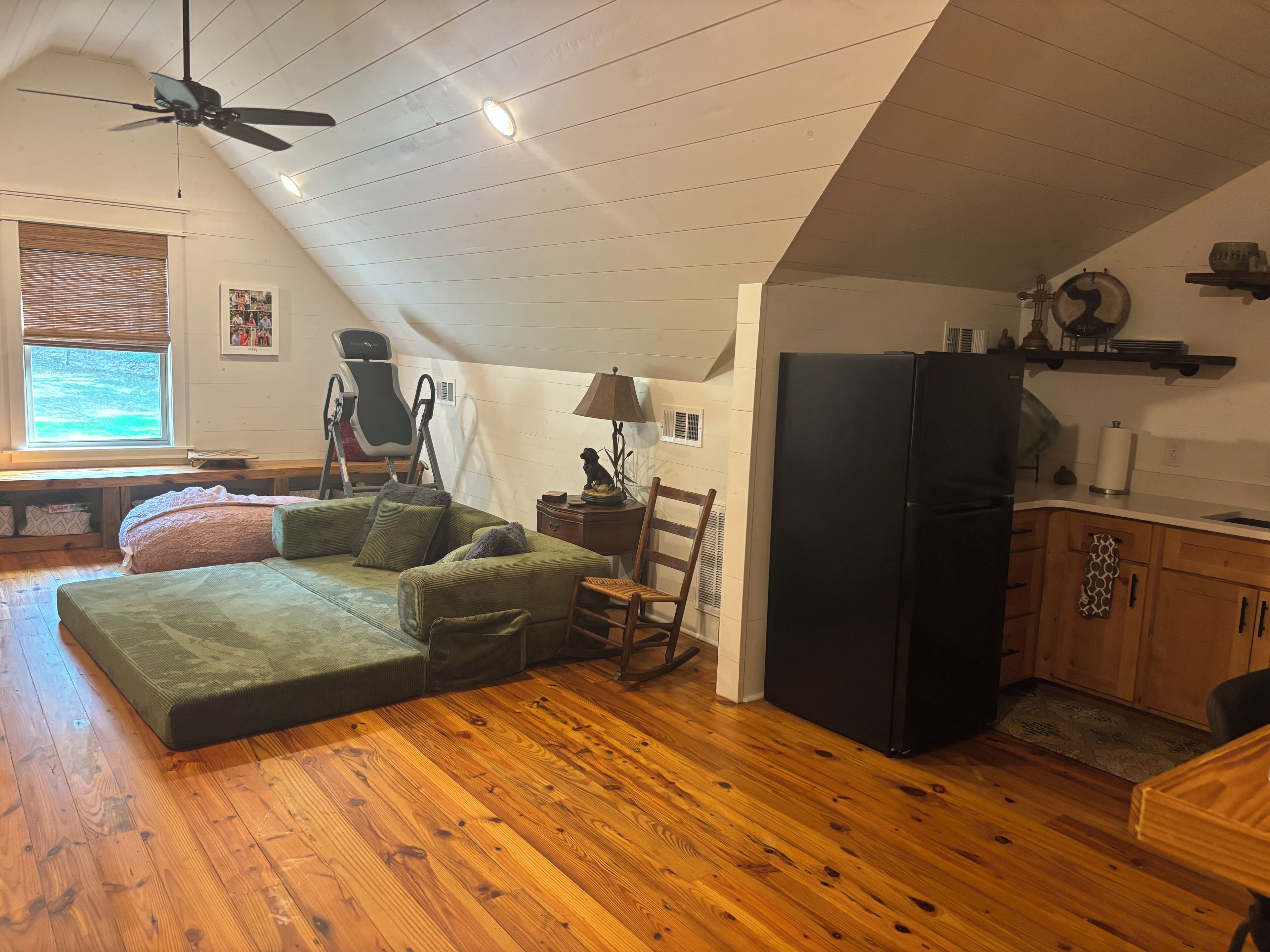 Un espace de vie de style loft avec un plancher en bois, un canapé d’angle vert, un fauteuil à bascule, un réfrigérateur noir et un coin cuisine doté de meubles en bois.