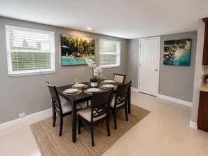 A dining area with a table set for four, surrounded by chairs, with a large window and a door in the background. - Casa Palma - A South Florida Oasis (Deerfield Beach)