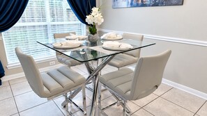 Uma área de refeições com mesa de vidro, quatro cadeiras e um vaso com flores brancas.