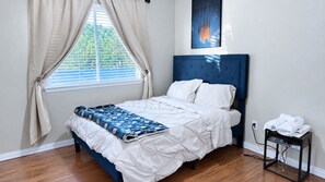 A bedroom with a bed, a window with curtains, a bedside table with towels, and a wall-mounted artwork.