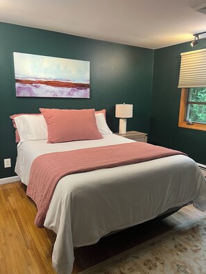 Een slaapkamer met een bed, nachtkastje, lamp, schilderij en een raam met jaloezieën.