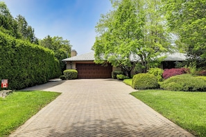 Une allée menant à une maison et à un garage, bordée de haies et d’arbres bien entretenus.