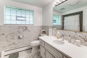 Une salle de bain équipée de toilettes, d’un lavabo, d’un miroir et d’une fenêtre avec des stores.