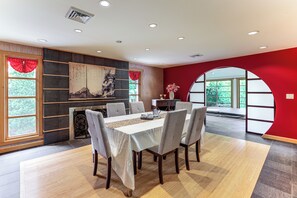 Un coin repas avec une table dressée pour six personnes, une cheminée et une grande fenêtre donnant sur un paysage verdoyant.