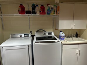 A laundry area with two white washing machines, a shelf with various cleaning products, and a sink with a bottle of bleach. - Your Lakefront Escape: 3BR Retreat with Japanese Garden and Sunset View (Lake View)