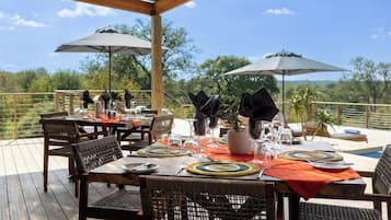 An outdoor dining area with tables set for two, surrounded by umbrellas, on a wooden deck with a view of the landscape.