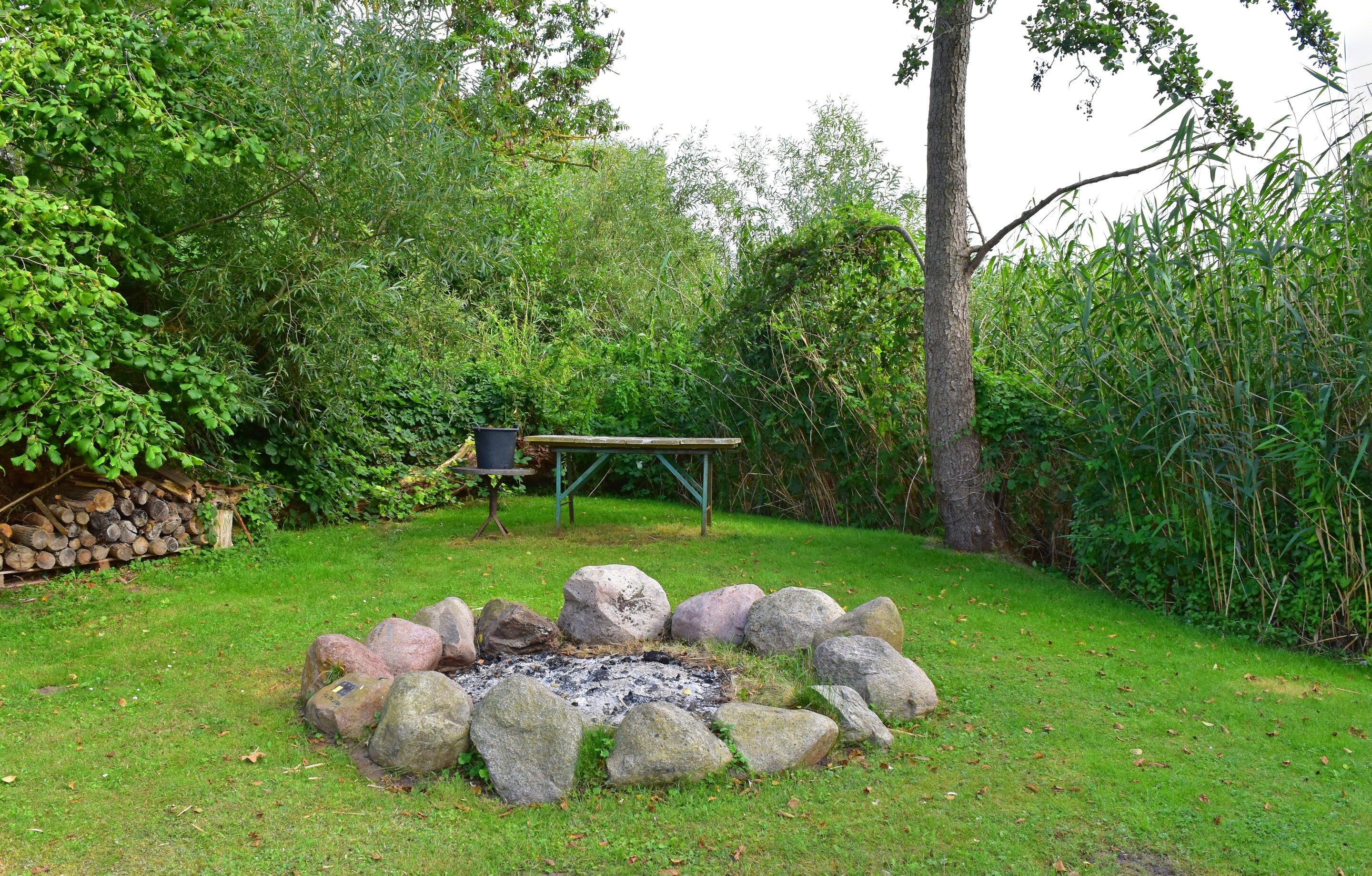 Un espace extérieur naturel avec un foyer entouré de rochers, un banc en bois et une pile de bois de chauffage.