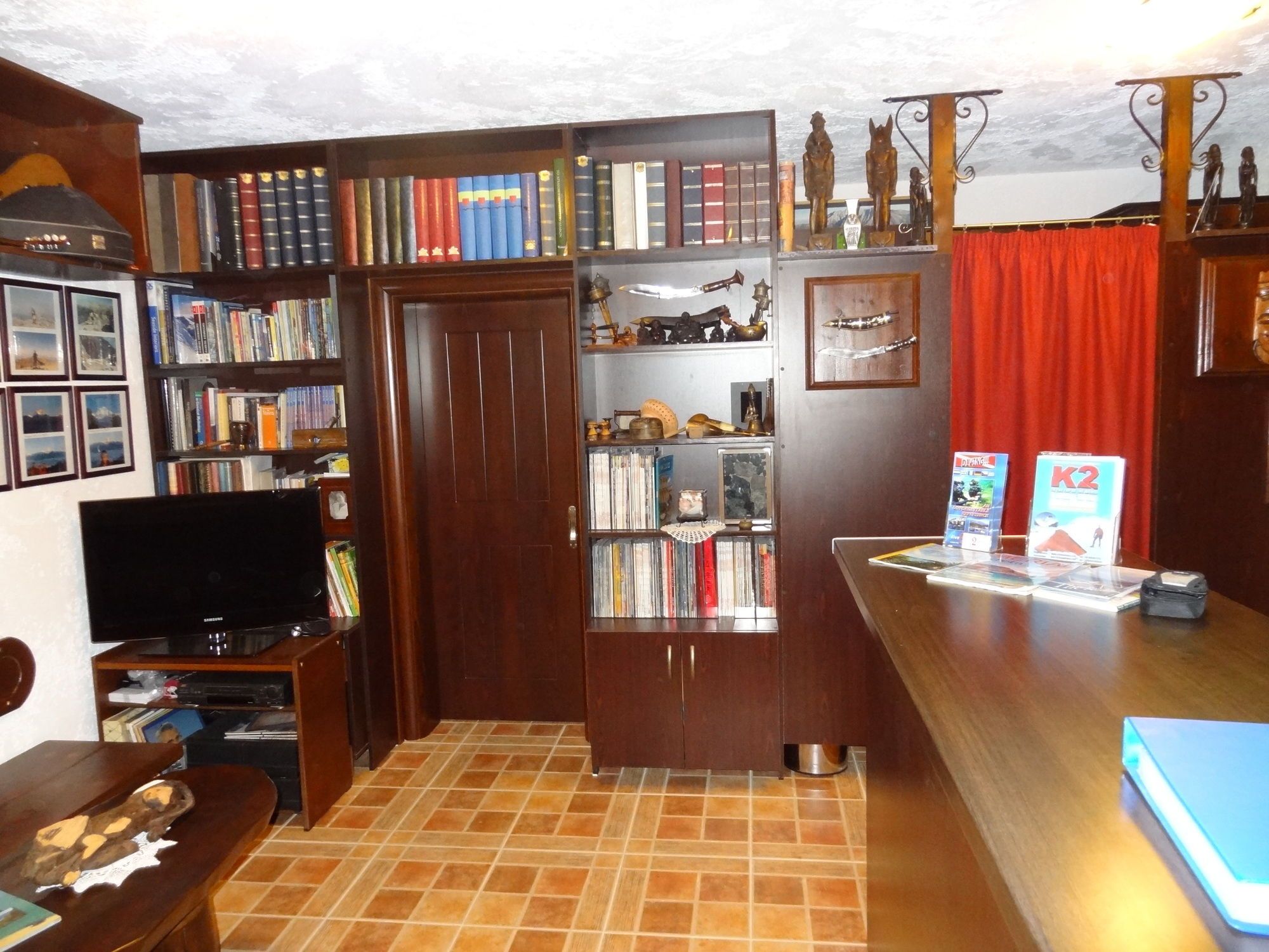 Ein Zimmer mit Fernseher, Bücherregalen, einem Schreibtisch mit Computer und einem roten Vorhang.