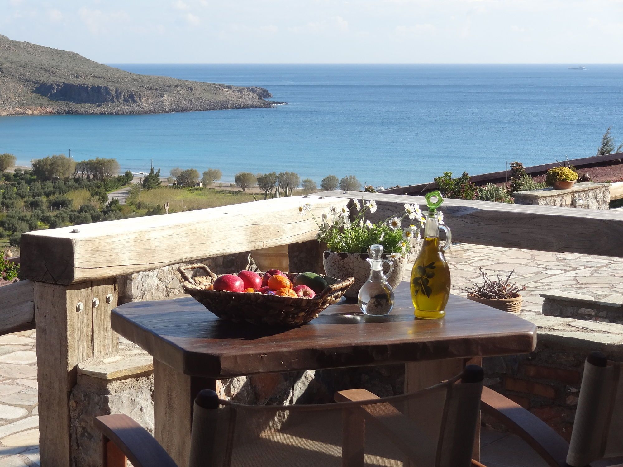 Un tavolo con una ciotola di frutta, una bottiglia e un vaso di fiori, con vista sulla costa.
